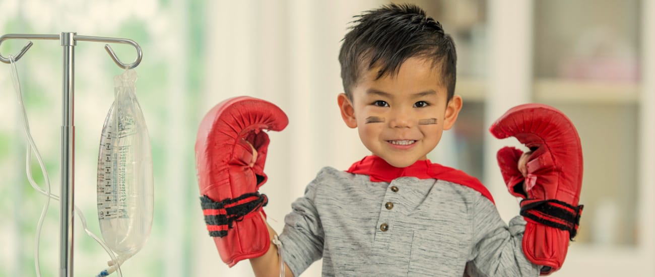 A little boy wearing boxing gloves stands next to a drip stand